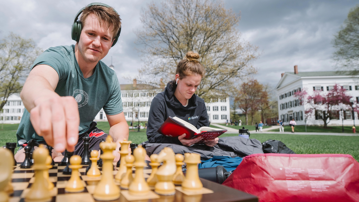 A photo of students spending time on the Green playing chess and reading a book