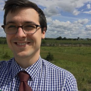 selfie of a young man with dark hair and glasses in front of a field