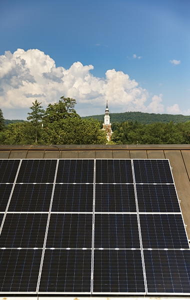 A photo of solar panels on the roof of a building on campus with Baker Tower in the background