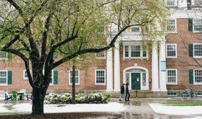 A photo of students walking on front of a building surrounded by grass and spring leaves covered in a thin layer of snow