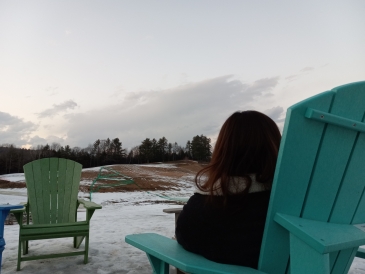 A girl with long brown hair shown from behind sitting on a blue chair looking at a field of melting snow.