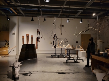 A woman with a mask is standing in an arts studio dimly lit with multiple art pieces made from wires hanging above. 