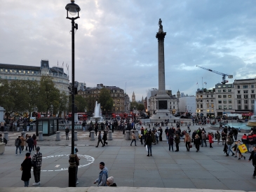 Trafalgar Square at sunset: Nelson's column at the center, buildings at the back, and people walking around