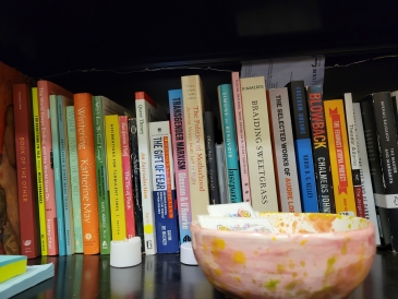 A bookshelf filled with a variety of colorful books on topics from feminism to ecology, accompanied by a bowl with paint splatters, and small items like sticky notes and a white cap.