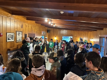 An aerial shot of around 40 people crowded in a log cabin. Someone at the front seems to be speaking, with everyone around listening in some kind of a circle.