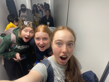 Three girls have their faces in surprised expressions for a selfie. They are boarding an airplane, with people and luggage in the background.