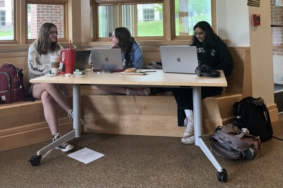 There are three people in the picture, all sitting around a table with their laptops open. Two of them are talking to each other, while the third looks at their phone. They all look very happy.