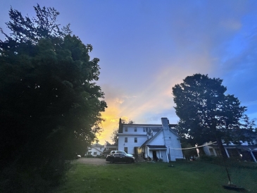 A three-story white house on the Dartmouth College campus. The photo is taken from the backyard in front of a volleyball net, with the sun setting in the background.