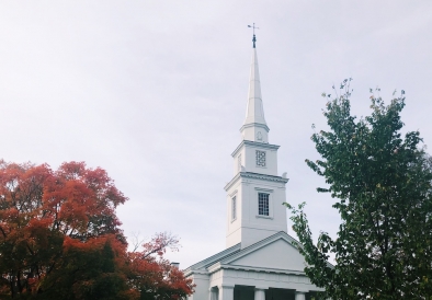 white church steeple against a cloudy sky with red and green leaves surrounding 