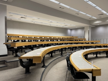 five rows of tables and chairs in a lecture hall with grey carpet and walls