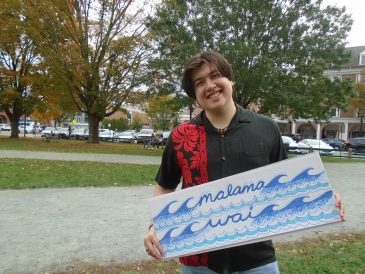 Gabriel Gilbert '23 standing with "Malama Wai" sign, dressed in black and red aloha shirt and light jeans