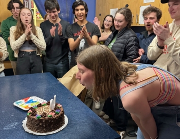 A photo of me blowing out the candles of a cake. The cake reads "Happy Birthday, Julia" and you can see friends behind me cheering. We are in a kitchen.