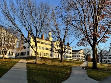 A photo of Dartmouth Hall during sunset. The building is light yellow with the glow from the sun, and barren trees surround the sidewalk in front of it. 