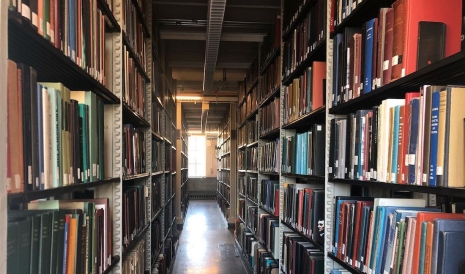 The stacks in Dartmouth's Baker library--shelves of books on either side and light coming in through a window at the end of the aisle.