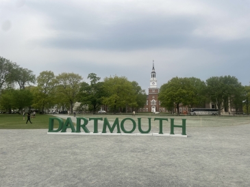 An image of a slightly cloudy day in the middle of the Dartmouth Green with giant green letter spelling out DARTMOUTH in front of Baker-Berry Library 