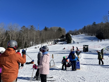 A photo of the Skiway. There are skiiers of all ages waiting at the bottom of the Magic Carpet, and there is a bright blue sky.