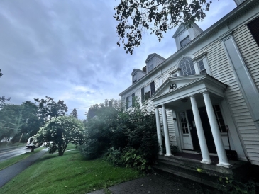 A white house with the letter A and Greek letter theta over the door, in front of a cloudy sky. Outside of the house to the left are green bushes and a tree with white flowers