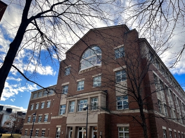 A picture of a large brick building. In the background, a bright blue, cloudy sky. In the foreground, tree branches. The building has a gigantic, semicircle window on the fourth floor.