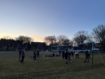 A photo of a volleyball net set up on the Green. The sun is setting in the background, and there are many students standing around the net playing volleyball.