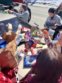 A photo of food on the trunk of a car. There are multiple students eating and making snacks around the trunk.