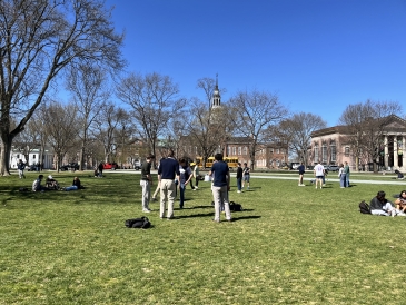 A photo of the Green on a sunny day. There is a group of students playing hacky sack, and other students lounging on the grass.