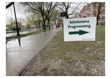 a picture of a yard sign with the text Admissions Programming Dartmouth with a large arrow pointing to the right in green text next to a sidewalk on a rainy day