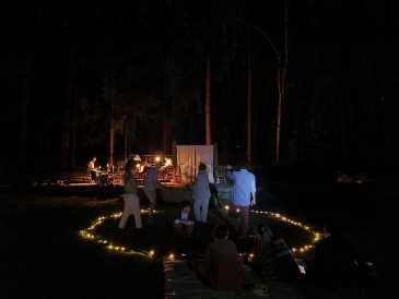 An outdoor night-time rehearsal with people gathered around a lit stage, surrounded by trees and illuminated by string lights on the ground, creating a cozy, intimate setting.