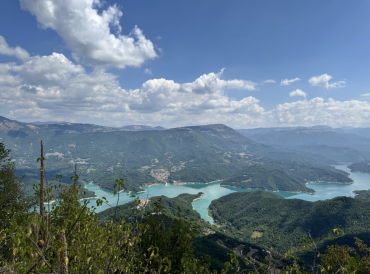 A panoramic view of a bright blue lake nestled in between green mountains. The sky is bright blue and full of clouds. 