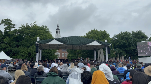  A large stage on the Green with Baker Tower in the background. In the foreground, there are many rows of people with raincoats on. The sky is cloudy and gray.