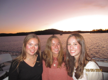 three girls are smiling on a boat. There is a sunset behind them.