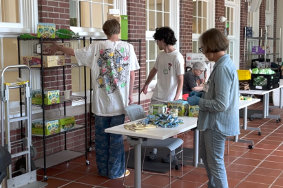 There are four people in the picture, all positioned around tables and shelves filled with granola bars. The room they are in is bright, and they appear to be sorting the food.