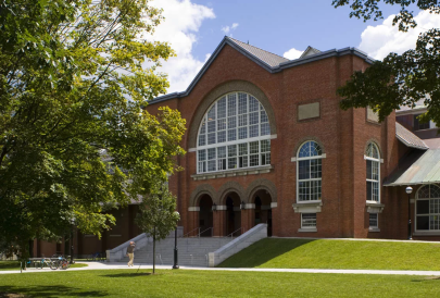 The exterior of the Alumni Gym facility, shadowed by large green trees and a vast grassy area borders the pavement. 