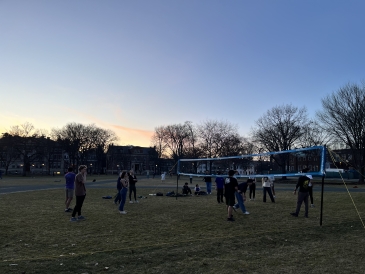  Students playing volleyball with a net on the Green. The sun has almost set, and there are many barren trees visible in the background.