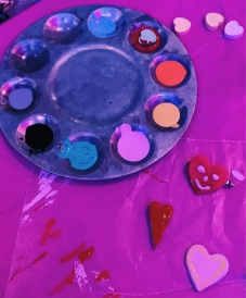 A paint palette, candy hearts, and painted clay hearts on top of a pink table clothe