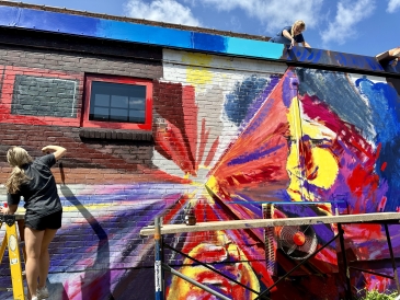 Two girls paint a brightly colored brick wall of someone blowing a dandelion of light.