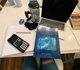 A notebook, textbook, laptop, calculator, and mask spread out over a desk.