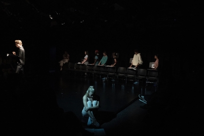 A picture of a theatre production taken inside of a black box theatre. It is dark, with a bright light shining on a man in the front corner, and a blue light on a girl sitting on the floor to his left