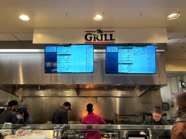 A grill station at a cafe with workers preparing food, featuring various cooking equipment and ingredients in the background.