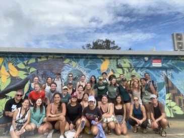 A group of about 30 students stand, smiling, in front of a mural on Waiheke Island in Auckland.
