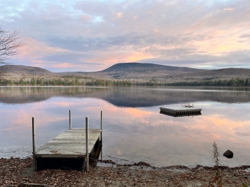 A photo of a dock in a lake. In the background, there is a mountain and a pink sunrise.