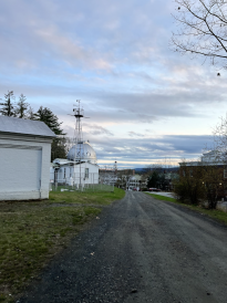 The road beside Shattuck Observatory just before sunset. There are many clouds in the pastel sky, and the trees in the background are barren. 