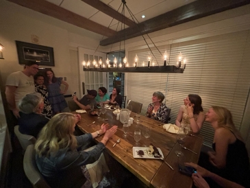 A crowded dinner table with a candle-style light fixture overhead. Three people stand in the far left corner, and everyone else at the table is cheering for them.