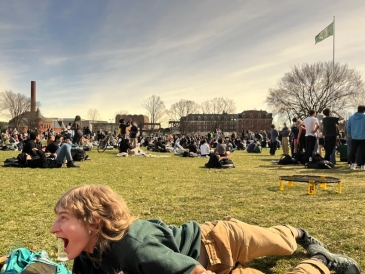  A crowd of people are sitting on the Green with their heads tilted towards the sky. The sky is dim. You can see campus buildings in the far back.