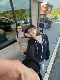 Two girls smile for a selfie next to a grocery store. One is wearing a tank top and the other is wearing a baseball cap and a zip-up hoodie. The girl in the tank top holds up a brown paper bag with groceries in it.