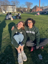 Two girls smile in Dartmouth sweatshirts while sitting on grass. The picture is taken on the Green, with some campus buildings in the background. There are other people sitting on the Green in the background, too. It looks sunny out.