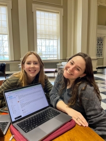 Two girls smile in a big room with gray walls. The girls are sitting down next to a laptop with its screen shown in the picture. The screen shows an email exchange.