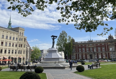 An image of a town square with a fountain and flowers, surrounded by buildings