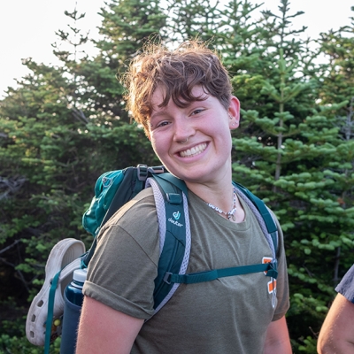 Headshot of Valen hiking Mt. Moosilauke. They are smiling widely and wearing a green t-shirt with a teal frame pack. 