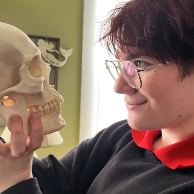 Lilla, a young person with dyed red hair, glasses, and a cool bird-themed sweatshirt, looks at a replica of a human skull