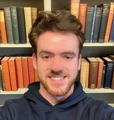 A selfie of a young man smiling. He has brown hair, a red beard, and is wearing a blue hoodie.